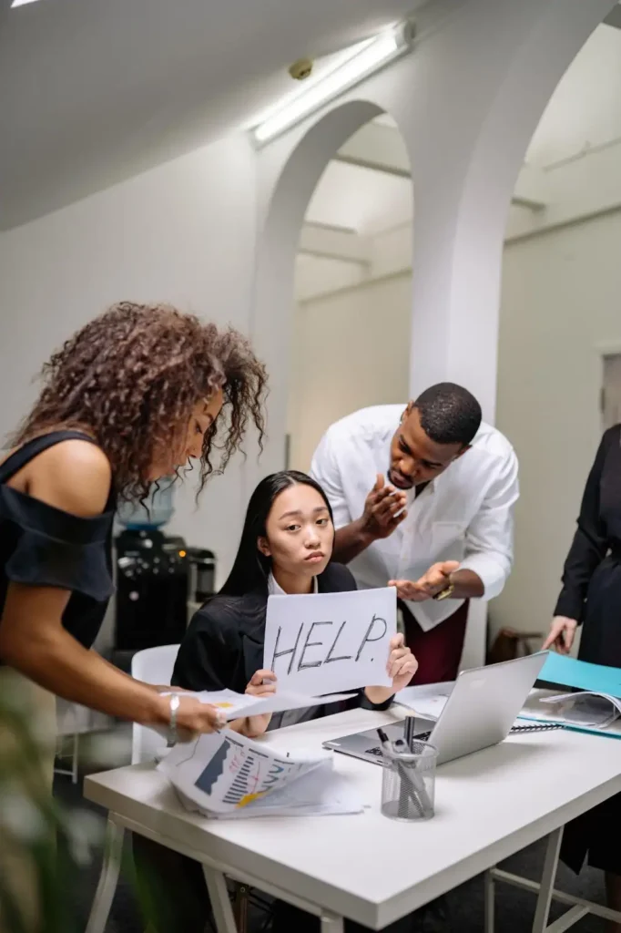 A woman sits at a desk holding a sign that says “HELP” while three colleagues stand around her, appearing to discuss documents and work.