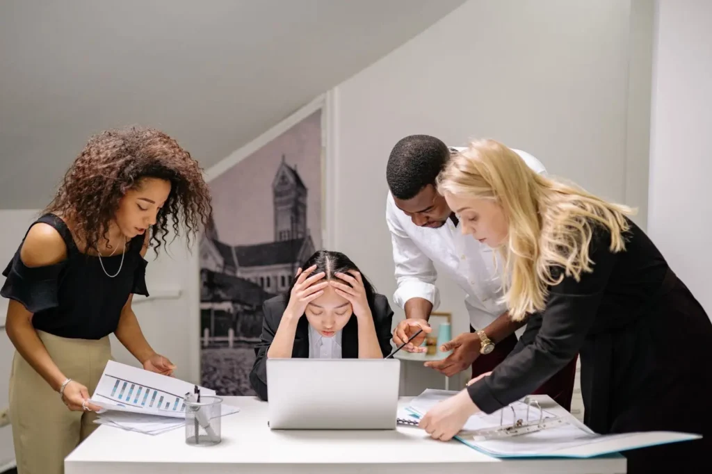 Four coworkers gather around a desk. One person looks stressed with hands on head, while others discuss documents and point at a laptop screen.