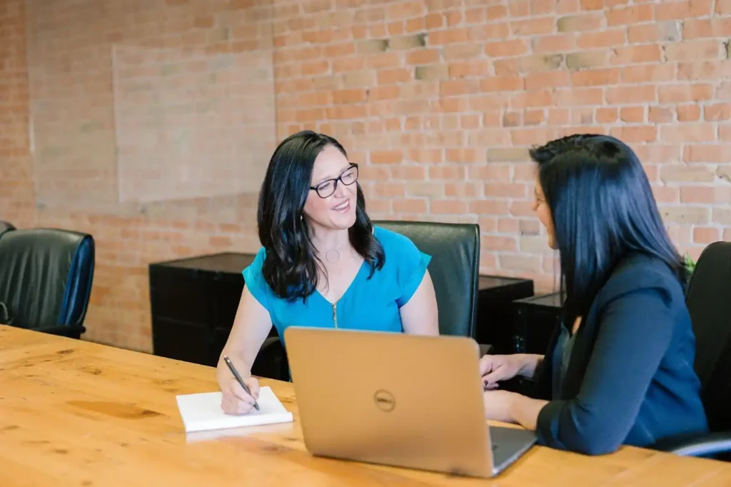 Two women sit at a conference table talking, perhaps discussing maternity policies or support. One is writing in a notebook, and a laptop is open in front of them. Both appear engaged in conversation in an office setting with a brick wall.