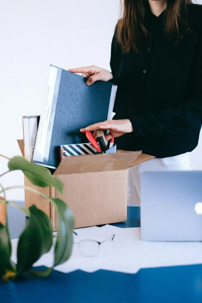 Person packing office supplies, folders, and notebooks into a cardboard box on a desk with a laptop, plant, and glasses nearby.
