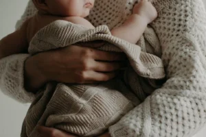 An adult in a knitted sweater tenderly cradles a baby wrapped in a beige knitted blanket, capturing a warm moment of maternity.