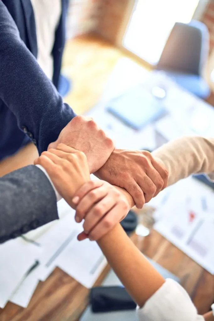 Four people standing around a table grasp each other’s wrists, forming a square shape, symbolizing teamwork or unity in a professional setting.