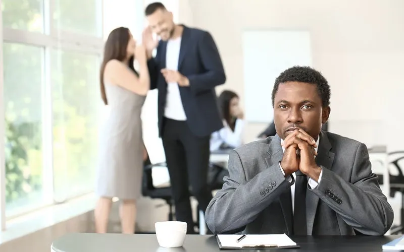 A man in a suit sits at his desk looking thoughtful, with a notebook and coffee cup in front of him—perhaps considering strategies used by racial discrimination lawyers. Three colleagues talk in the blurred background of a modern office.