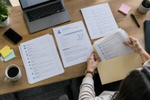 Person reviews resumes at a desk with a laptop, coffee, and handwritten notes—searching for retaliation evidence among printed documents to help prove retaliation in the workplace.